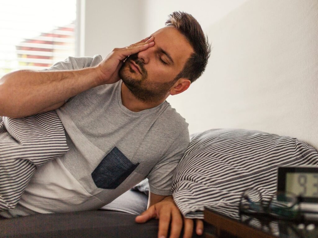 Homem acorda cansado com a mão no rosto, deitado na cama. O relógio digital ao lado marca 9:30, refletindo a exaustão matinal apesar das horas de sono.
