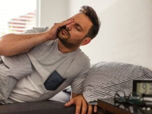 Homem acorda cansado com a mão no rosto, deitado na cama. O relógio digital ao lado marca 9:30, refletindo a exaustão matinal apesar das horas de sono.